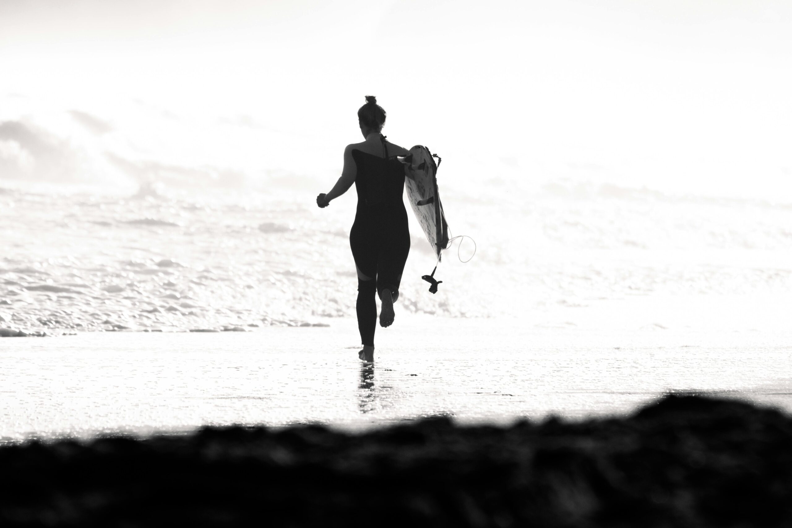 A woman carrying a surfboard runs along the beach, captured in black and white.